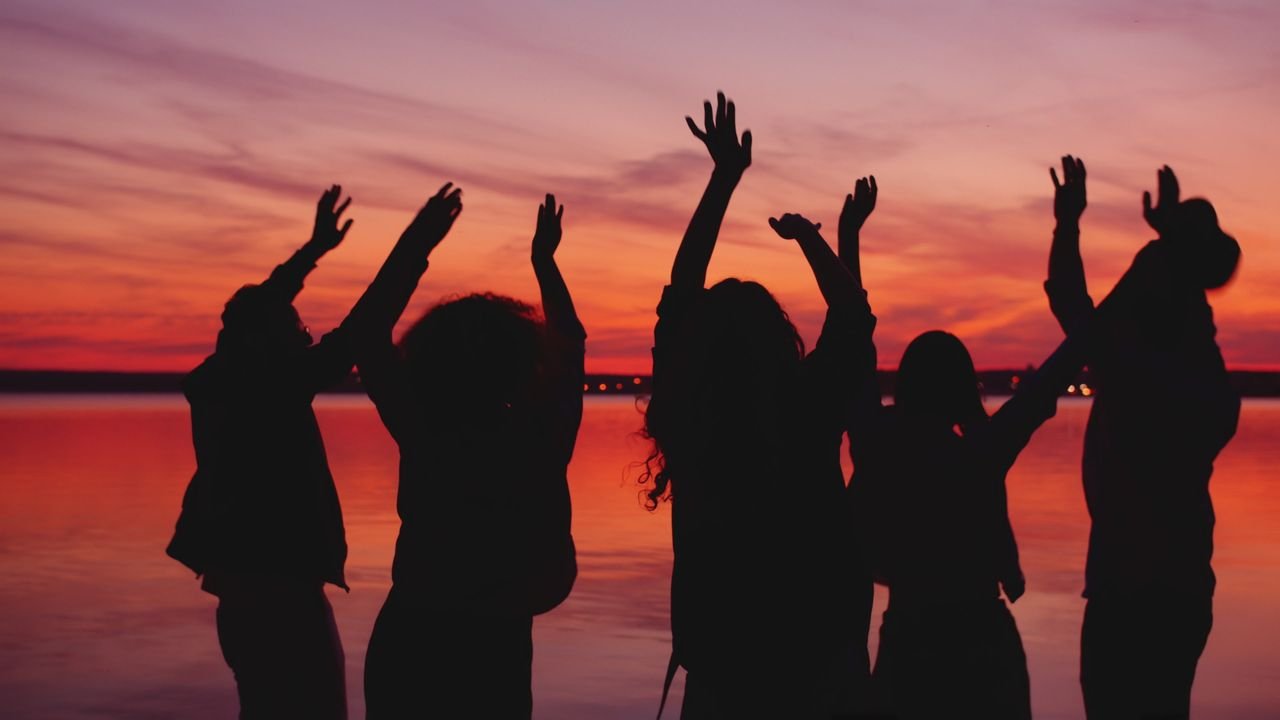 A group of people silhouetted against a vibrant sunset near a body of water, expressing joy.