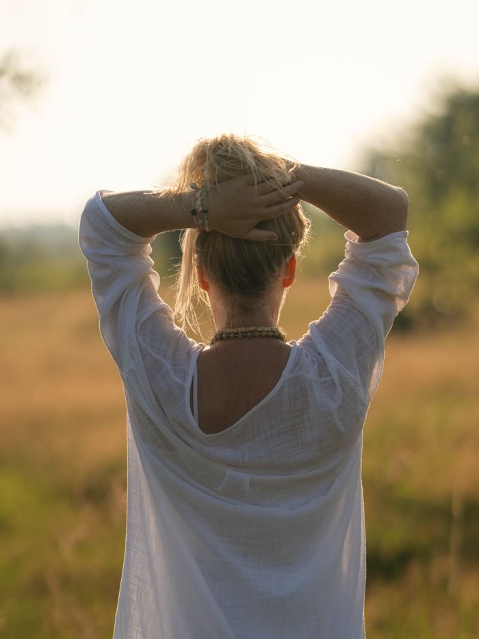 Rear view of a woman in a white shirt with hands up, enjoying a sunny day outdoors.